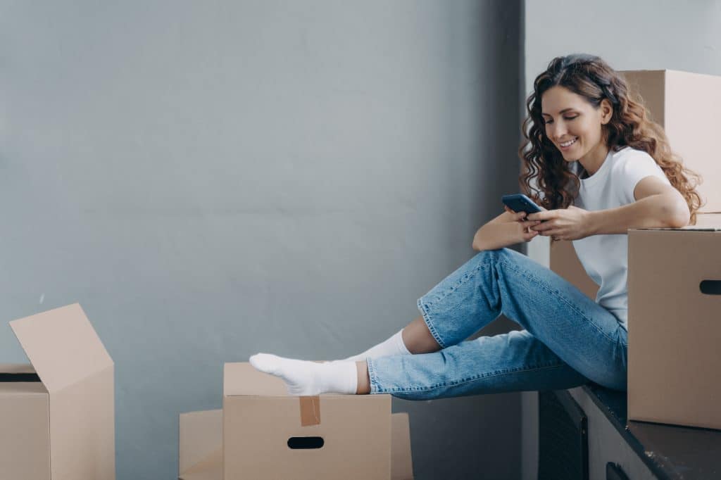 Woman sitting atop moving boxes leaving a moving company review on her phone