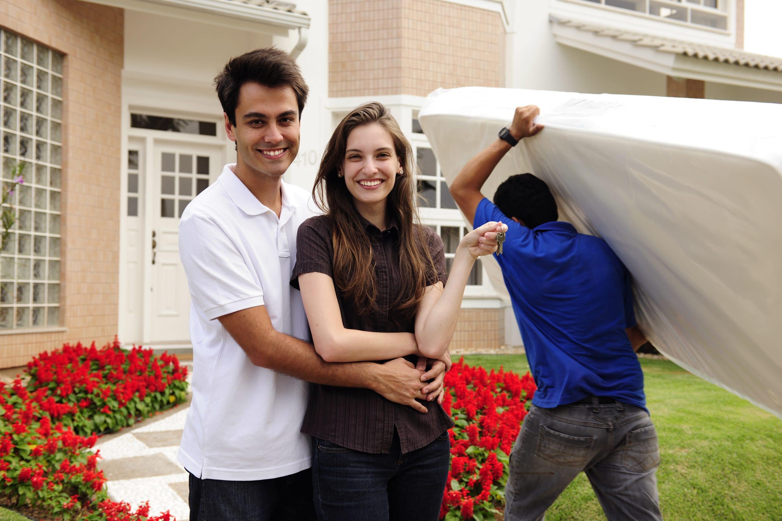 Couple showing keys and relocator carrying mattress into their new home