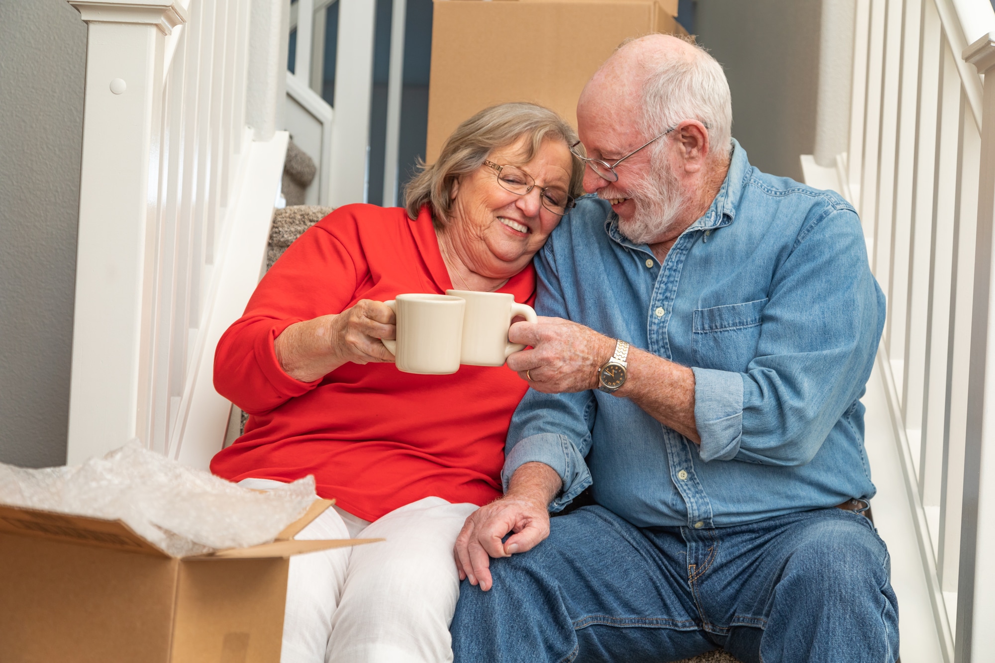 senior couple toasting each other with mugs of coffee. There are moving boxes around them